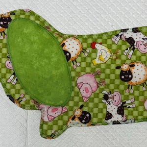 May include: A Dachshund-shaped fabric item with a green checkered pattern and farm animal illustrations. The design includes cows, sheep, chickens, and pigs. A large green oval shape is on the dog's back, and a smaller green shape is on the tail.