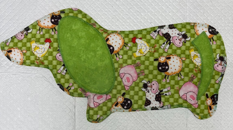 May include: A Dachshund-shaped fabric item with a green checkered pattern and farm animal illustrations. The design includes cows, sheep, chickens, and pigs. A large green oval shape is on the dog's back, and a smaller green shape is on the tail.