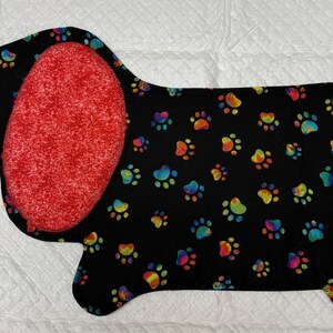 May include: A black dachshund-shaped mat with a colorful paw print pattern. The ears and tail are red. The mat is likely for a pet, possibly a dog.
