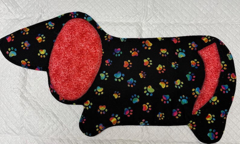 May include: A black dachshund-shaped mat with a colorful paw print pattern. The ears and tail are red. The mat is likely for a pet, possibly a dog.
