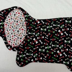 May include: A dog-shaped fabric item with a black background covered in white bones, red bows, and green accents. The dog's ear features a white background with paw prints in red, brown, and gray. The item is likely a placemat or pet bed.