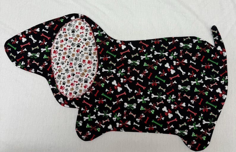 May include: A dog-shaped fabric item with a black background covered in white bones, red bows, and green accents. The dog's ear features a white background with paw prints in red, brown, and gray. The item is likely a placemat or pet bed.