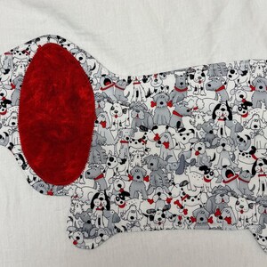 May include: A dog-shaped fabric applique featuring a pattern of various dog breeds in gray, black, and white. The dog's ear is a solid, textured red. The applique is likely for crafting or sewing projects.