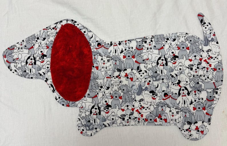 May include: A dog-shaped fabric applique featuring a pattern of various dog breeds in gray, black, and white. The dog's ear is a solid, textured red. The applique is likely for crafting or sewing projects.