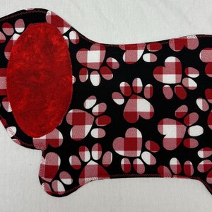 May include: A Dachshund-shaped fabric item with a black background and red and white paw prints. The dog's ear is solid red. The edges are stitched with black thread. The design is reminiscent of a dog bed or placemat.