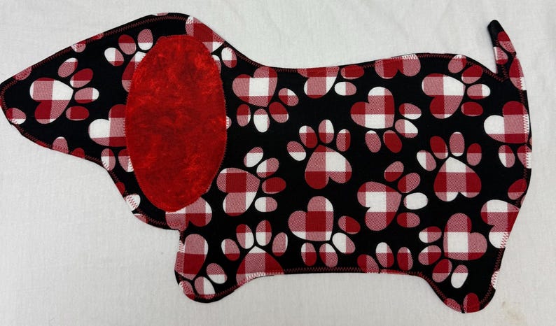 May include: A Dachshund-shaped fabric item with a black background and red and white paw prints. The dog's ear is solid red. The edges are stitched with black thread. The design is reminiscent of a dog bed or placemat.