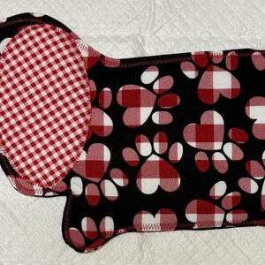 May include: A dog-shaped pet bed with a black background and red and white paw prints. The ears and a section of the body are red and white checkered fabric. The bed has a black stitched edge.