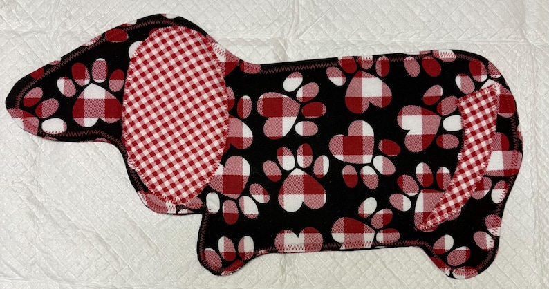 May include: A dog-shaped pet bed with a black background and red and white paw prints. The ears and a section of the body are red and white checkered fabric. The bed has a black stitched edge.