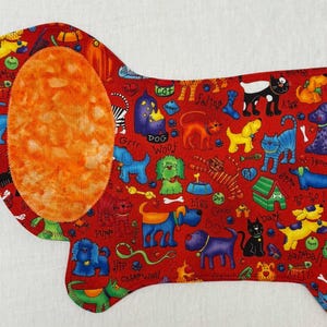 May include: A red fabric dog-shaped placemat with a large orange ear. The placemat is covered in colorful cartoon dogs, bones, and dog-related phrases. The background is white.