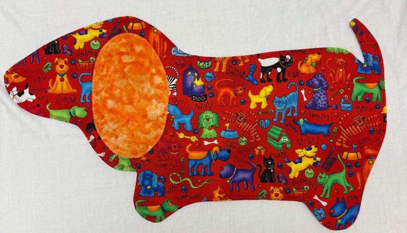 May include: A red fabric dog-shaped placemat with a large orange ear. The placemat is covered in colorful cartoon dogs, bones, and dog-related phrases. The background is white.