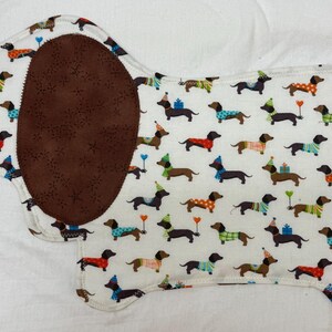 May include: A dachshund-shaped fabric item with a white background covered in repeating dachshund illustrations. The dachshunds wear party hats and sweaters. A large brown oval and a brown stripe are on the item.