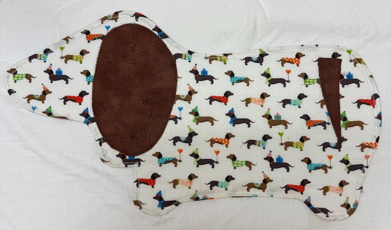 May include: A dachshund-shaped fabric item with a white background covered in repeating dachshund illustrations. The dachshunds wear party hats and sweaters. A large brown oval and a brown stripe are on the item.