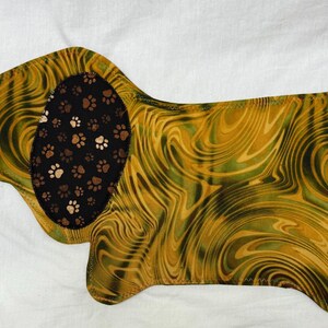 May include: A dog-shaped fabric item with a swirling pattern in shades of gold and green. The dog's ear is black with brown and white paw prints. The item is likely a placemat or decorative piece.