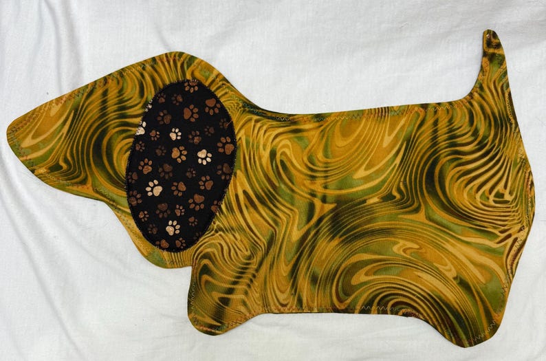 May include: A dog-shaped fabric item with a swirling pattern in shades of gold and green. The dog's ear is black with brown and white paw prints. The item is likely a placemat or decorative piece.