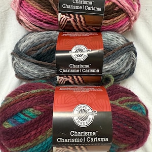 以下が含まれることがあります： さまざまな色のLoops & Threads Charisma糸の3つのかせ。上のかせはピンク、茶色、バラ色です。真ん中のかせはグレーと茶色です。下のかせは、バーガンディ、ティール、グリーンです。各かせにはブランド名が記載されたラベルが付いています。