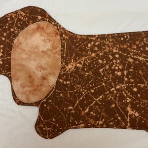 May include: A brown dachshund-shaped fabric item with a speckled pattern and a light brown ear and tail detail. The fabric has a splattered design in white and light brown, creating a unique, artistic look. The item is likely a decorative piece.