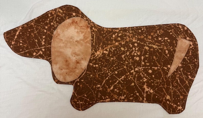 May include: A brown dachshund-shaped fabric item with a speckled pattern and a light brown ear and tail detail. The fabric has a splattered design in white and light brown, creating a unique, artistic look. The item is likely a decorative piece.