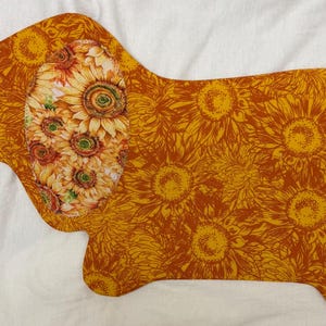 May include: A dog-shaped placemat featuring a sunflower design. The dog's ear is adorned with a cluster of sunflowers, while the body is covered in a pattern of yellow sunflowers on a brown background. The placemat is on a white surface.