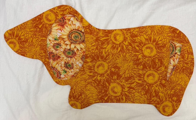 May include: A dog-shaped placemat featuring a sunflower design. The dog's ear is adorned with a cluster of sunflowers, while the body is covered in a pattern of yellow sunflowers on a brown background. The placemat is on a white surface.