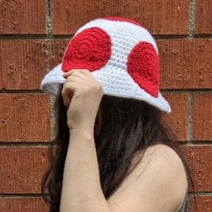 May include: White crochet bucket hat with red mushroom-shaped appliques. The hat has a wide brim and a rounded crown.