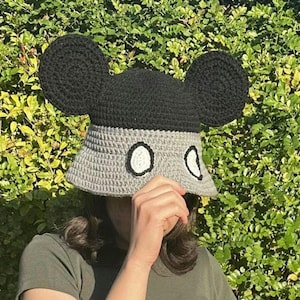 May include: A gray and black crocheted bucket hat with large black ears, resembling Mickey Mouse. The hat has white circles on the front.