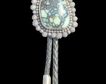 May include: A bolo tie with a large, oval turquoise stone set in a silver-colored frame. The stone has a mottled pattern of blue and green. The frame is surrounded by a decorative border. The tie has a braided cord and silver-colored tips.