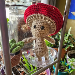 May include: A crocheted mushroom doll with a red cap and brown body. The doll is sitting on a shelf with other plants.
