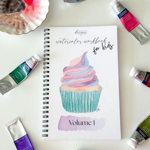 May include: A watercolor workbook for kids, featuring a cupcake illustration, surrounded by tubes of watercolor paint. The workbook is spiral-bound and labeled "Volume 1". Paint colors include blue, pink, purple, green, and orange.