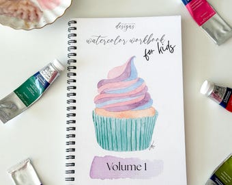 May include: A watercolor workbook for kids, featuring a cupcake illustration, surrounded by tubes of watercolor paint. The workbook is spiral-bound and labeled "Volume 1". Paint colors include blue, pink, purple, green, and orange.