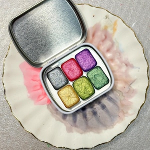 May include: A small, open, silver metal tin containing six rectangular watercolor paint pans in various colors: silver, red, purple, green, yellow, and gold. The tin sits on a white plate with a gold rim and watercolor paint splotches.
