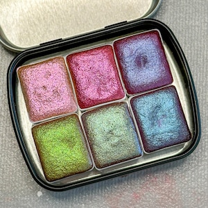 May include: A metal tin with six watercolor paint pans. The pans are filled with shimmery, iridescent paint in shades of pink, green, blue, and purple.