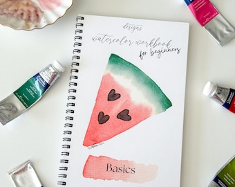 May include: A spiral-bound watercolor workbook for beginners featuring a painted watermelon slice with heart-shaped seeds and the text "designs watercolor workbook for beginners" and "Basics," surrounded by several tubes of watercolor paint.
