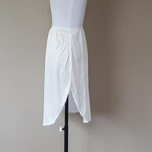 May include: A white half slip, displayed on a mannequin. The slip has a straight cut with a slit on the side and a scalloped hem. The fabric appears to be lightweight and slightly textured.