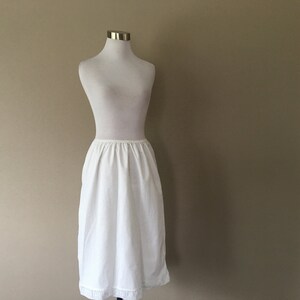 May include: A white cotton slip with a lace trim. The slip is on a white mannequin and has a gathered waist.