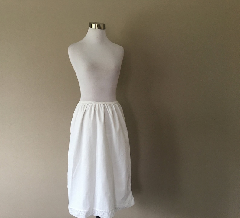 May include: A white cotton slip with a lace trim. The slip is on a white mannequin and has a gathered waist.