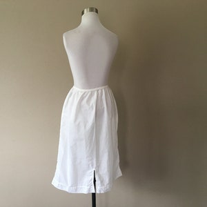 May include: A white cotton slip skirt with a straight silhouette and a back slit. The skirt is on a white mannequin.