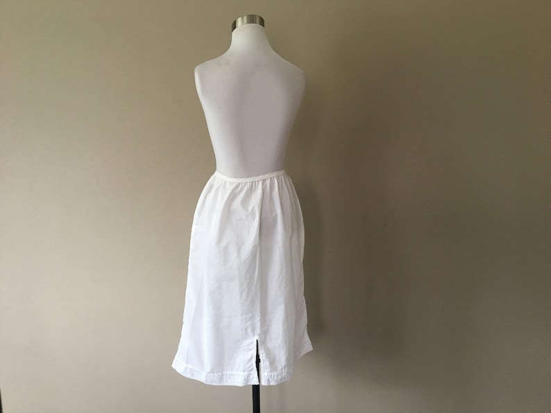 May include: A white cotton slip skirt with a straight silhouette and a back slit. The skirt is on a white mannequin.
