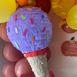 May include: A large, ice cream cone-shaped pi&ntilde;ata. The top is a lavender sphere with colorful rectangular confetti. The cone is made of cream-colored fringed paper. Balloons in shades of yellow, orange, and brown are in the background.