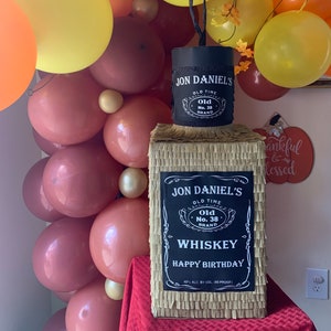 K&ouml;nnte beinhalten: Eine rechteckige Pi&ntilde;ata in Form einer Whiskeyflasche, bedeckt mit beigefarbenem Fransenpapier. Sie hat ein schwarzes Etikett mit wei&szlig;em Text: JON DANIEL'S OLD TIME OLD NO. 38 BRAND WHISKEY HAPPY BIRTHDAY 40% ALC. BY VOL. (80 PROOF).
