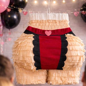 May include: A piñata shaped like a lower torso wearing beige fringed paper, a red fabric panel with black scalloped edges, and a small pink heart accent at the waistband. The piñata is suspended by a white string against a party background.