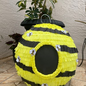 May include: A round, yellow and black bee-themed pi&ntilde;ata with a large circular opening. The pi&ntilde;ata is decorated with black stripes and small bee stickers. A white metal handle is attached to the top.