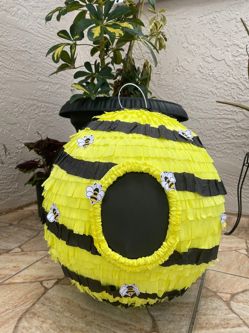 May include: A round, yellow and black bee-themed pi&ntilde;ata with a large circular opening. The pi&ntilde;ata is decorated with black stripes and small bee stickers. A white metal handle is attached to the top.