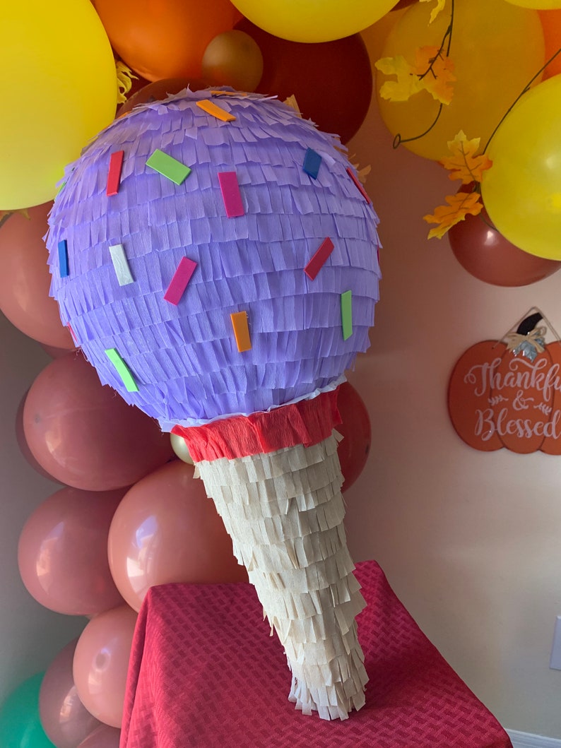 May include: A large, ice cream cone-shaped pi&ntilde;ata. The top is a lavender sphere with colorful rectangular confetti. The cone is made of cream-colored fringed paper. Balloons in shades of yellow, orange, and brown are in the background.