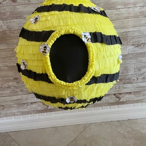May include: A spherical pi&ntilde;ata designed to resemble a beehive. The pi&ntilde;ata is yellow with black stripes and decorative bee stickers. It has a large, round opening and a white string for hanging.