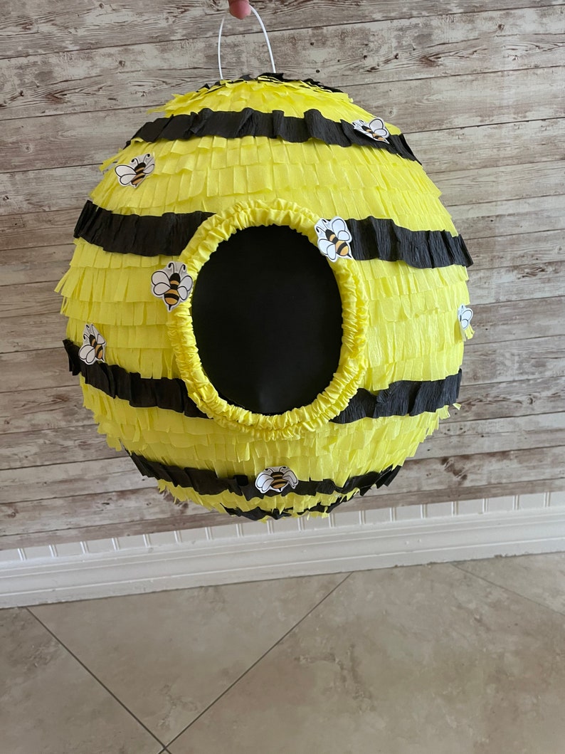 May include: A spherical pi&ntilde;ata designed to resemble a beehive. The pi&ntilde;ata is yellow with black stripes and decorative bee stickers. It has a large, round opening and a white string for hanging.