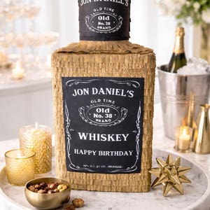 K&ouml;nnte beinhalten: Eine rechteckige Pi&ntilde;ata in Form einer Whiskeyflasche mit goldenen Fransen und einem schwarzen Etikett mit der Aufschrift JON DANIEL'S OLD TIME OLD NO. 38 BRAND WHISKEY HAPPY BIRTHDAY. Der Flaschenhals ist schwarz und tr&auml;gt ein passendes Etikett.