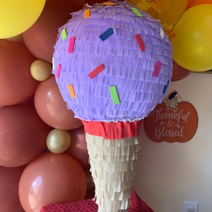 May include: A large, handmade ice cream cone pi&ntilde;ata. The cone is made of cream-colored paper fringe, topped with a lavender scoop decorated with colorful confetti. The pi&ntilde;ata is set against a backdrop of balloons in shades of yellow, peach, and brown.