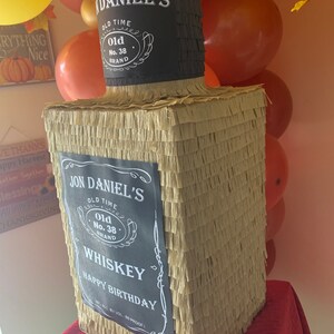 K&ouml;nnte beinhalten: Eine rechteckige Pi&ntilde;ata in Form einer Whiskeyflasche, bedeckt mit beigem und schwarzem Fransenpapier. Das schwarze Etikett tr&auml;gt den Text JON DANIEL'S OLD TIME Old No. 38 BRAND WHISKEY HAPPY BIRTHDAY. Der Flaschenhals ist schwarz mit Etikett.
