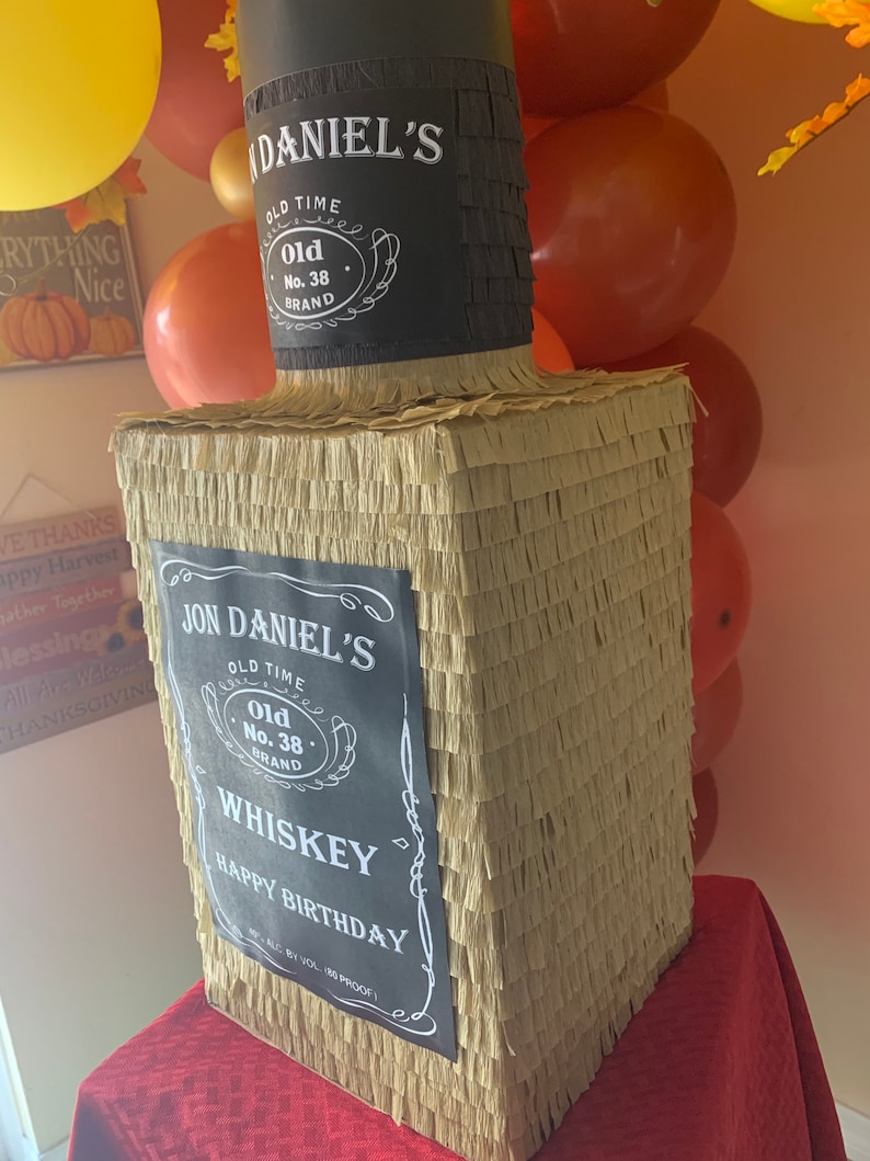 K&ouml;nnte beinhalten: Eine rechteckige Pi&ntilde;ata in Form einer Whiskeyflasche, bedeckt mit beigem und schwarzem Fransenpapier. Das schwarze Etikett tr&auml;gt den Text JON DANIEL'S OLD TIME Old No. 38 BRAND WHISKEY HAPPY BIRTHDAY. Der Flaschenhals ist schwarz mit Etikett.