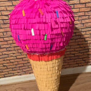 May include: A large, handmade pi&ntilde;ata shaped like an ice cream cone. The top is a vibrant pink, decorated with colorful confetti-like accents. The cone is a light tan color, with a textured, layered design. A festive party decoration.